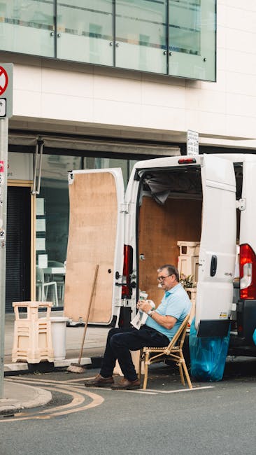 A man wearing a black cap, white T-shirt, and blue jeans sits on the pavement beside the open rear doors of a large white van loaded with numerous cardboard boxes of various sizes, some sealed with pa