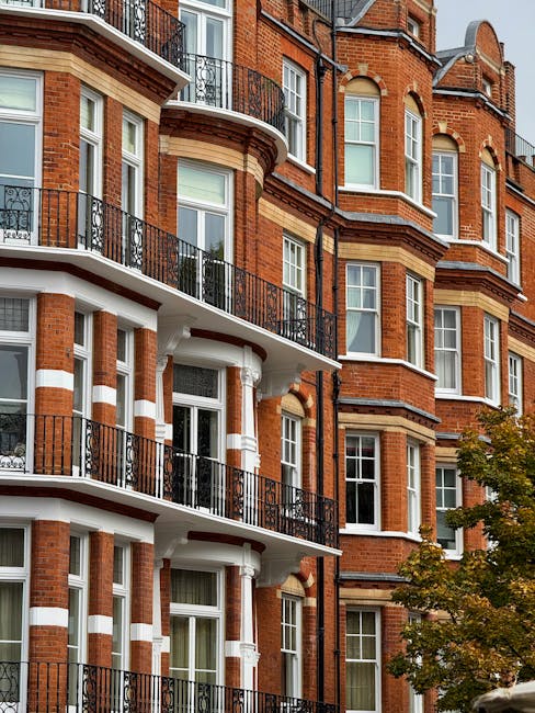 A narrow, cobbled street in a residential area with a slight curve, lined with multi-story terraced houses featuring white and cream facades, some with black metal balconies and window frames. Potted