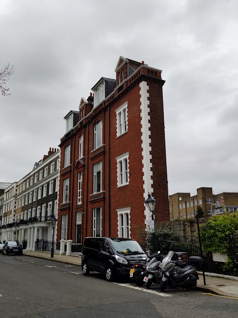 A residential street in South Kensington lined with parked cars on both sides, with several trees in full blossom displaying pink flowers along the pavement. The street is flanked by elegant Victorian